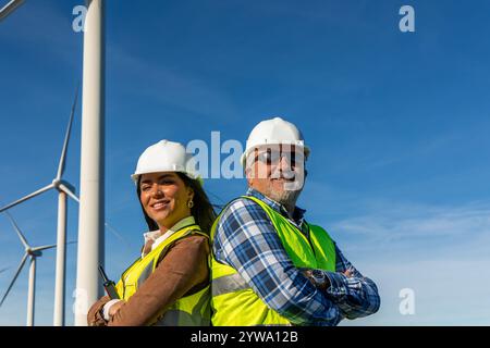 Two multiethnic engineers, a man and a woman, posing back to back with crossed arms, wearing safety helmets and vests in a wind turbine power plant un Stock Photo