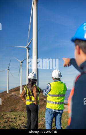 Three multiethnic engineers wearing safety vests and helmets are analyzing a wind turbine farm, contributing to sustainable energy development and a g Stock Photo