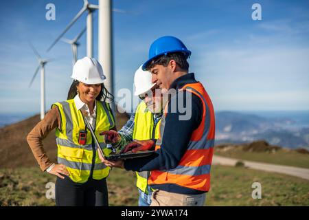 Three multiethnic engineers wearing safety vests and helmets are using a laptop in a wind turbine field, collaborating on a project related to renewab Stock Photo