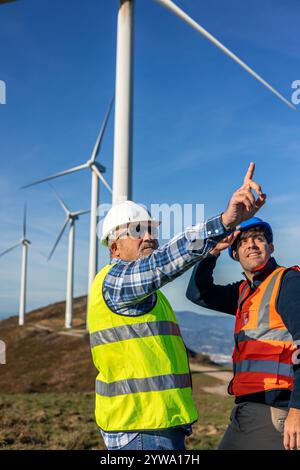 Two engineers in safety vests and helmets discussing a wind turbine at a wind farm on a sunny day, highlighting the growth of green energy and sustain Stock Photo
