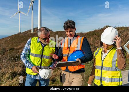 Three multiethnic engineers wearing safety vests are discussing a wind turbine maintenance project using a digital tablet in a field, promoting renewa Stock Photo