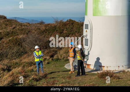 Three engineers wearing safety vests and hard hats are discussing next to the base of a wind turbine, contributing to clean energy generation Stock Photo