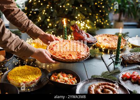 Man places Christmas quiche pie on festive table surrounded by many dishes in cozy evening atmosphere with Christmas tree and garlands Stock Photo