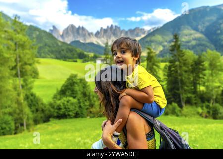 Mother carrying her son on her shoulders while enjoying the view of the odle mountains and the funes valley in the dolomites in summer Stock Photo