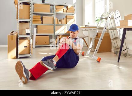 Male worker storekeeper in overall sitting on the floor in warehouse after accident. Stock Photo