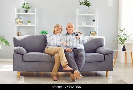 Senior couple using remote control to change channel watching TV sitting on sofa at home together. Stock Photo