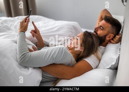 Young happy loving couple is lying in bed in the morning. They are relaxing and using smartphone. Stock Photo