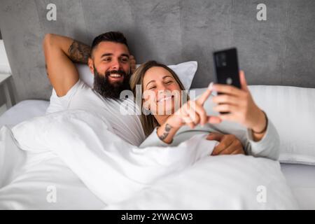 Young happy loving couple is lying in bed in the morning. They are relaxing and using smartphone. Stock Photo