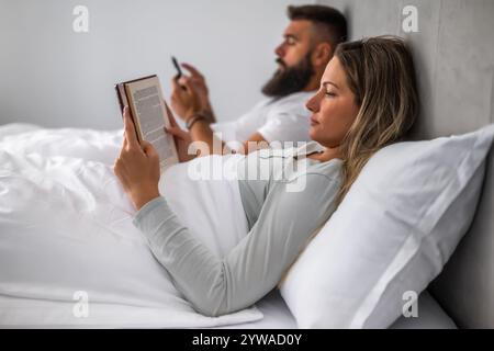 Young couple is lying in bed in the evening. Woman is reading book and man is using smartphone. Stock Photo