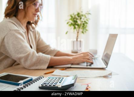 Beautiful middle-aged woman in glasses making marks in financial documents at home office using laptop. Small business, home finances, money savings c Stock Photo