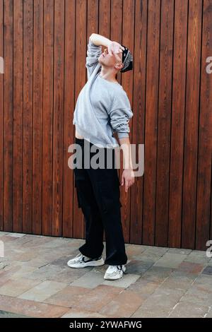 A person in a grey sweatshirt and black pants is striking a pose in front of a wooden wall Stock Photo