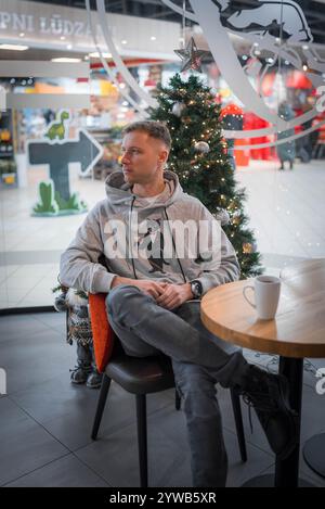 A young man in a gray hoodie with a cat graphic sits in a cafe in Lodz, Poland. A small Christmas tree and a warm beverage create a cozy atmosphere. Stock Photo