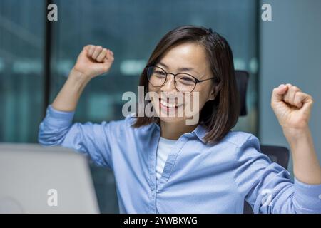 An Asian businesswoman joyfully raises her fists in celebration, highlighting her success at work. Her expression radiates happiness and achievement in a modern office Stock Photo