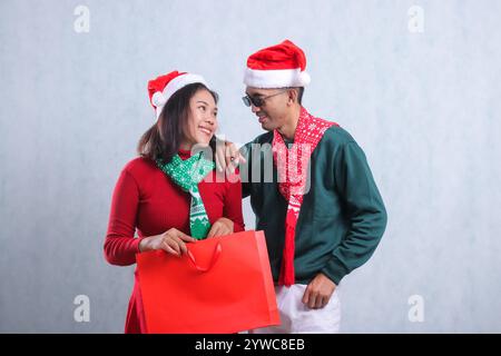 two asian man and woman wearing christmas sweaters, santa hats and scarves, happily looking at each other faces while carrying christmas gift paper ba Stock Photo