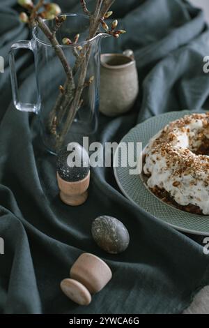 Close-up of a bundt cake with icing and chopped nuts, painted Easter eggs and a vase with cherry blossom branches on fabric Stock Photo