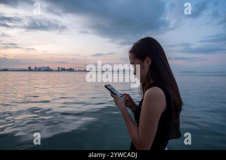 Side view of a young woman standing by the sea using her mobile phone, Tan Jetty, George Town, Penang, Malaysia Stock Photo