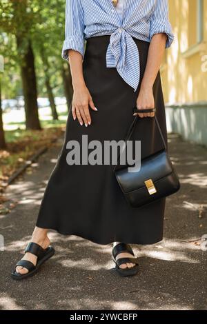 Neck down close-up of a stylish young woman in a black silk skirt, striped shirt and sandals standing in a city street in summer, Belarus Stock Photo