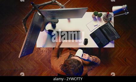 Top down view of self employed man doing IT support job from apartment personal office, coding on PC, drinking coffee. Software developer writing lines of code on computer at home, aerial shot Stock Photo