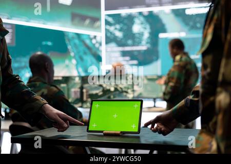 Two servicemen working in team with a green screen mockup, using a tablet to help troopers in power base command center. Soldiers examine real time information on device. Close up. Stock Photo