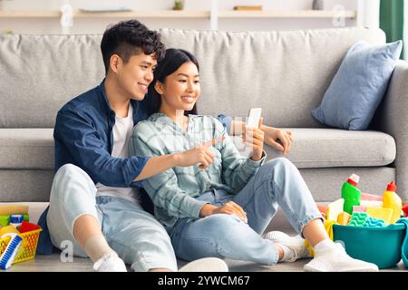 Loving chinese couple with cleaning tools resting, using smartphone Stock Photo