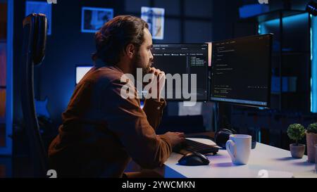 Programmer doing web development during remote job shift in neon lit home office, finishing coding project. IT specialist sitting at computer desk, working on desktop PC, camera B Stock Photo