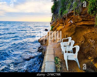 Three empty chairs sit on a ledge overlooking the ocean. The backdrop is an ochre-colored cliff wall. The sea is relatively calm and the sky is clear. Stock Photo