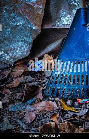A forgotten blue plastic leaf rake lies partially buried and leaning against a dirty, crinkled metallic silver tarp surrounded by damp Stock Photo