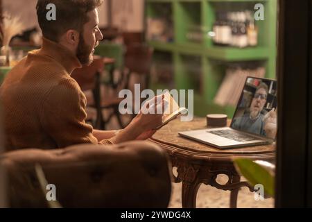 Young bearded man in orange turtleneck sweater having video call while taking notes in cozy cafe interior. Remote worker multitasking during virtual m Stock Photo