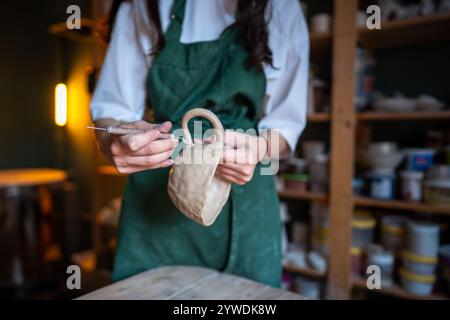 Girl ceramist in apron shaping cup handle, making clay mug during ceramics masterclass, hand closeup Stock Photo