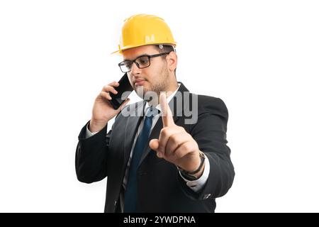 Busy male contractor or engineer wearing yellow helmet making wait gesture with index finger while speaking on smart phone isolated on white studio ba Stock Photo