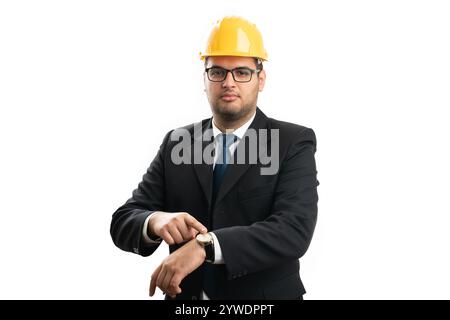 Serious male contractor architect wearing office suit and yellow helmet pointing index finger at wrist watch as time keeping concept isolated on white Stock Photo