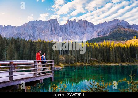 In the heart of the Dolomites, two hikers stand on a wooden deck, captivated by the turquoise lake and impressive mountain peaks. Lake Carezza or Karersee Dolomites in Italy at Autumn Stock Photo