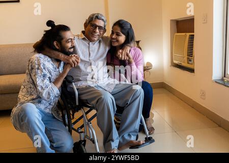 Happy Indian young couple spending time with senior disabled father on wheel chair at home. Happy parents Day. Stock Photo