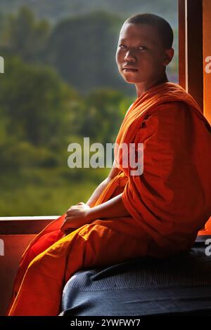 Young buddhist monk sitting next to the window and looks at me in Luang Prabang, Laos Stock Photo