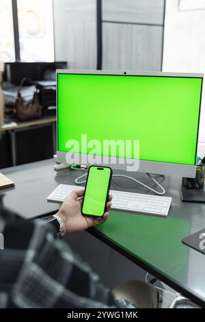 Office worker holding a smartphone with a vibrant green screen, positioned near a computer featuring a matching green screen on the desk Stock Photo