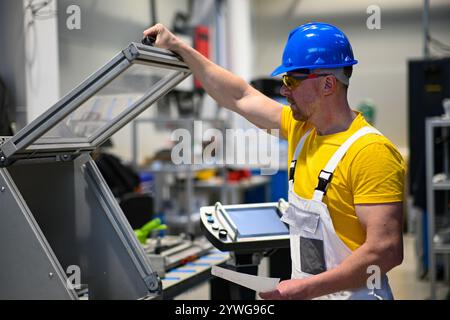 Factory worker wearing safety glasses and hardhat operating industrial saw and reading instructions. Stock Photo
