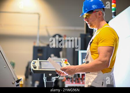 Factory worker wearing safety glasses and hardhat operating industrial saw and reading instructions. Stock Photo