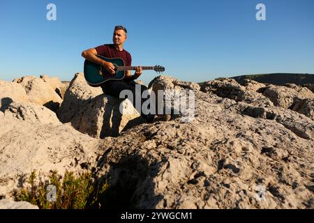A man in a dark t-shirt playing acoustic guitar on a rocky area by the sea Stock Photo