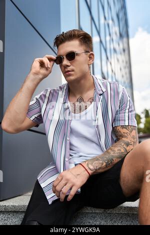 Young man with tattoos relaxes outside a contemporary structure on a sunny day. Stock Photo