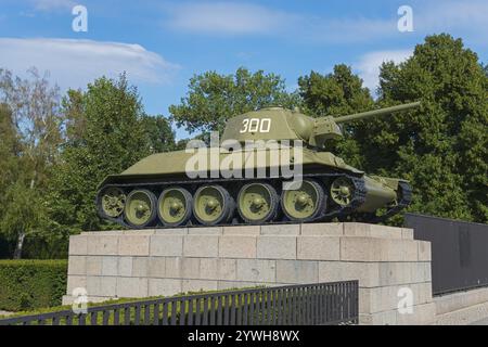 A green tank stands on a stone platform surrounded by trees and a blue cloudy sky in a park, Russian tank t-34, Soviet Memorial, Berlin, Tiergarten di Stock Photo