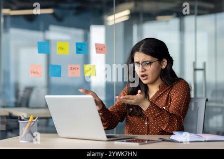 A businesswoman reacts with frustration using a laptop at workplace, conveying confusion and challenge. Modern office setting highlights a professional atmosphere and the realities of work stress. Stock Photo