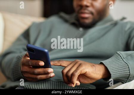 Close up of African American man holding phone in hand and tapping screen with finger while relaxing on couch at home, copy space Stock Photo