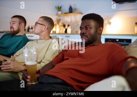 Side view portrait of young African American man sitting on couch with and watching TV with drinks at home Stock Photo