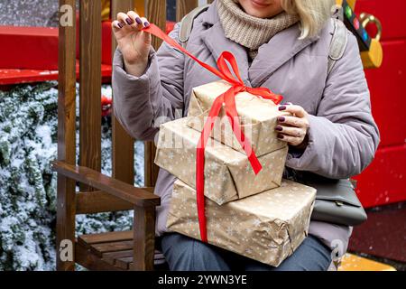 Woman's hands untie red ribbon on Christmas gifts wrapped in brown paper. Winter snowy day outside. Close-up. Stock Photo