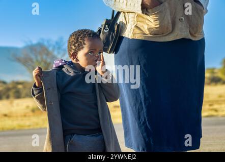 village, portrait african kid girl, going to school kindergarten with her mother, outdoors walking on the road, hand fingers in the mouth Stock Photo