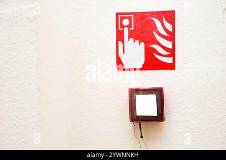 Fire alarm button encased in glass over a sign showing a finger pushing the button near flames on a white wall Stock Photo