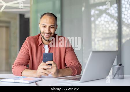 A cheerful male professional interacts with his smartphone while multitasking, working at his laptop. His positive demeanor embodies efficiency, communication, and productivity in work environment. Stock Photo