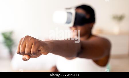 Virtual training. Overweight black lady in VR glasses using fighting simulator, making punch at home, focus on fist Stock Photo
