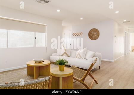 Modern living room with white sofa, light wood furniture, and beige rug. Large window, white walls, and wall hangings. Stock Photo