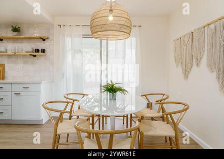 A bright, modern dining area features a white round table surrounded by wicker chairs. A large woven pendant light hangs above. Stock Photo